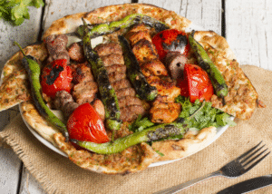 A plate filled with mixed grilled kebabs, including skewered beef and chicken, served on flatbread with charred green peppers, roasted tomatoes, and fresh herbs. The dish is placed on a rustic wooden surface with cutlery beside it.