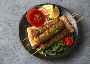 A plate of grilled kebab skewers served on flatbread with fresh parsley, sliced cherry tomatoes, red onion, lemon wedges, and a bowl of red dipping sauce. The meal is presented on a dark plate against a textured gray background.