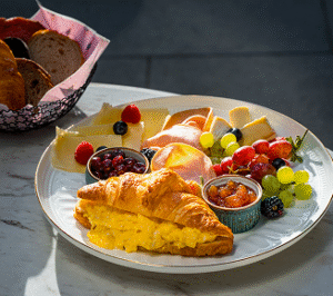 Plate of Mediterranean breakfast featuring a croissant stuffed with scrambled eggs, assorted cheeses, sliced turkey, fresh grapes, mixed berries, and fruit preserves on a white plate.
